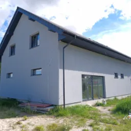 Nowoczesny dom jednorodzinny po remoncie elewacji, z szarą fasadą, ciemnym dachem i orynnowaniem, widoczny z perspektywy trawnika z chwastami i piaskiem.