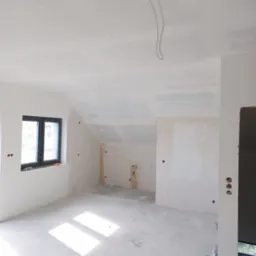 Wnętrze w trakcie remontu: ściany z płyt karton-gips, gładzie szpachlowe na ścianach i suficie, widoczne kable wystające z sufitu, okno z czarną ramą wpadające światło słoneczne na podłogę.