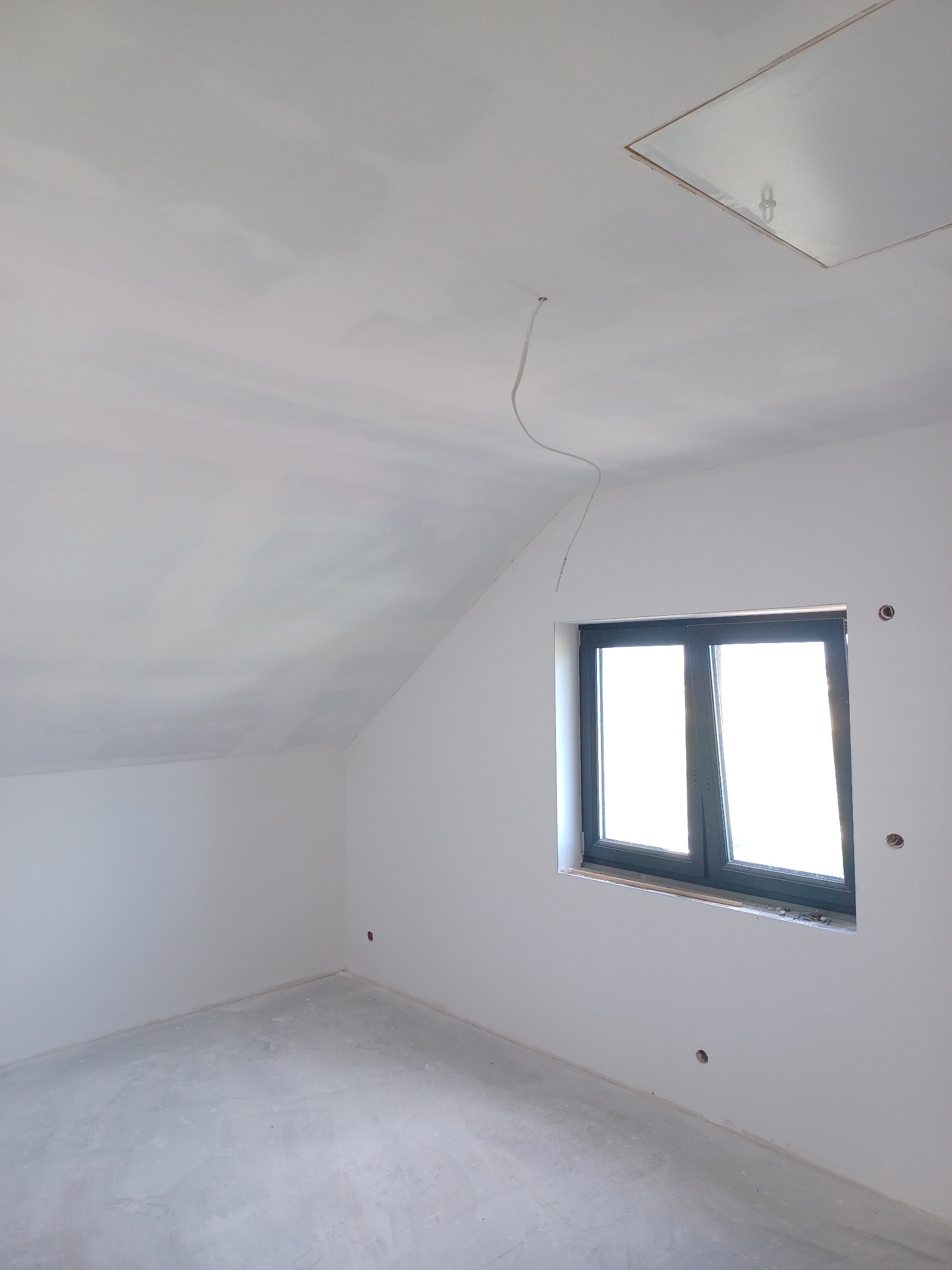 Poddasze po pracach wykończeniowych: ściany i sufit pokryte gładzią, widoczne otwory na gniazdka elektryczne, okno w ciemnej ramie i klapa włazu na strych.