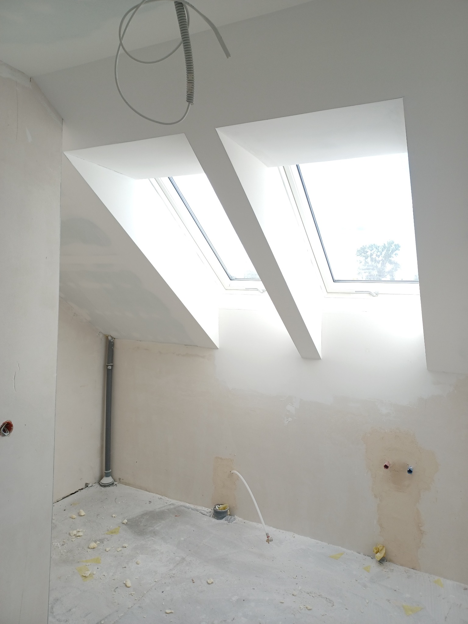 Poddasze w trakcie remontu: surowe ściany przygotowane pod gładzie, widoczne dwa okna dachowe, rury instalacyjne i resztki materiałów budowlanych na podłodze.