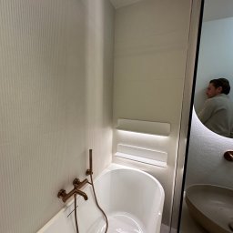 CORE PARTNER Bartłomiej Rejdych - Nowoczesna łazienka z białą wanną, złotą armaturą i podświetlanymi półkami, odbicie osoby w lustrze.