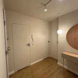 CORE PARTNER Bartłomiej Rejdych - Nowoczesny przedpokój z drewnianą podłogą, białymi drzwiami ukrytymi w ścianie, okrągłym lustrem w miedzianej ramie i minimalistyczną konsolą z jasnego drewna, oświetlony reflektorami i wiszącą...