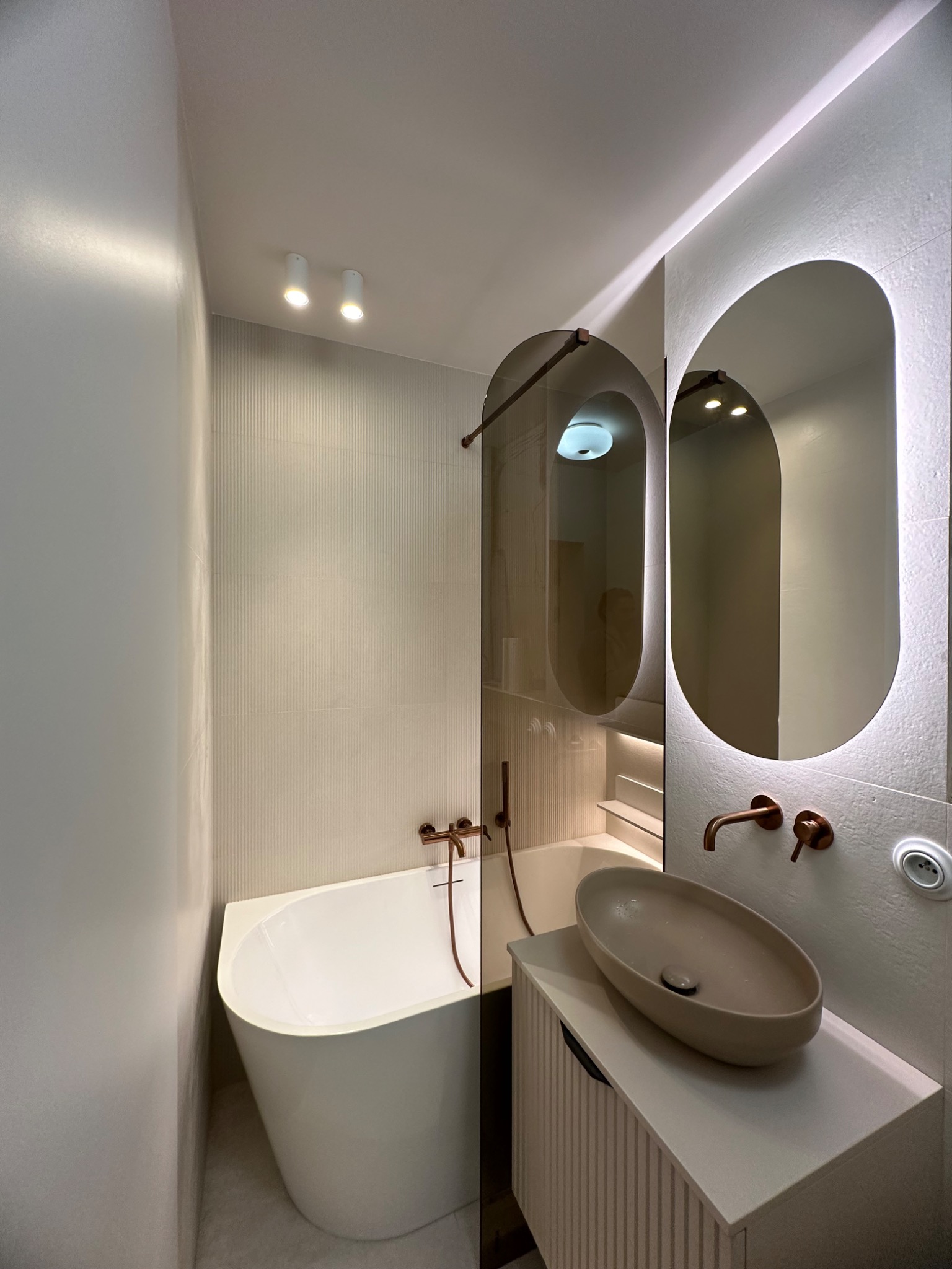Nowoczesna łazienka z owalnym lustrem podświetlanym LED, beżową umywalką nablatową i baterią w kolorze miedzi, minimalistyczna wanna i pionowe płytki na ścianie.
