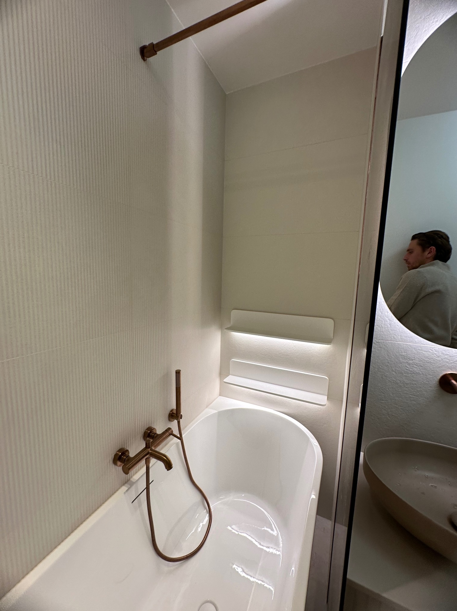 Nowoczesna łazienka z białą wanną, złotą armaturą i podświetlanymi półkami, odbicie osoby w lustrze.