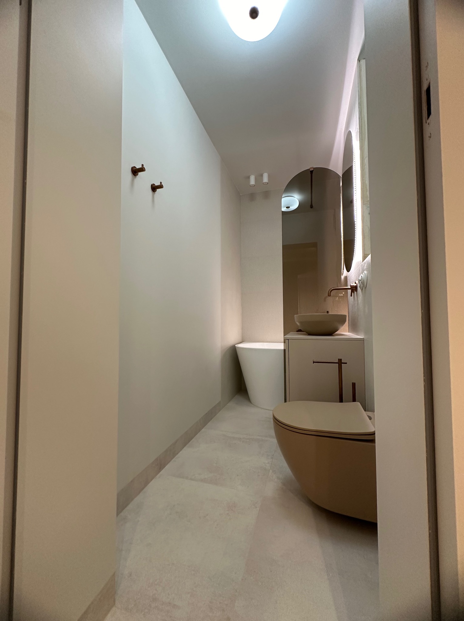 Nowoczesna łazienka w neutralnych kolorach z wiszącą toaletą w kolorze beżowym, umywalką nablatową i podświetlanym lustrem owalnym, widok zza futryny drzwi.