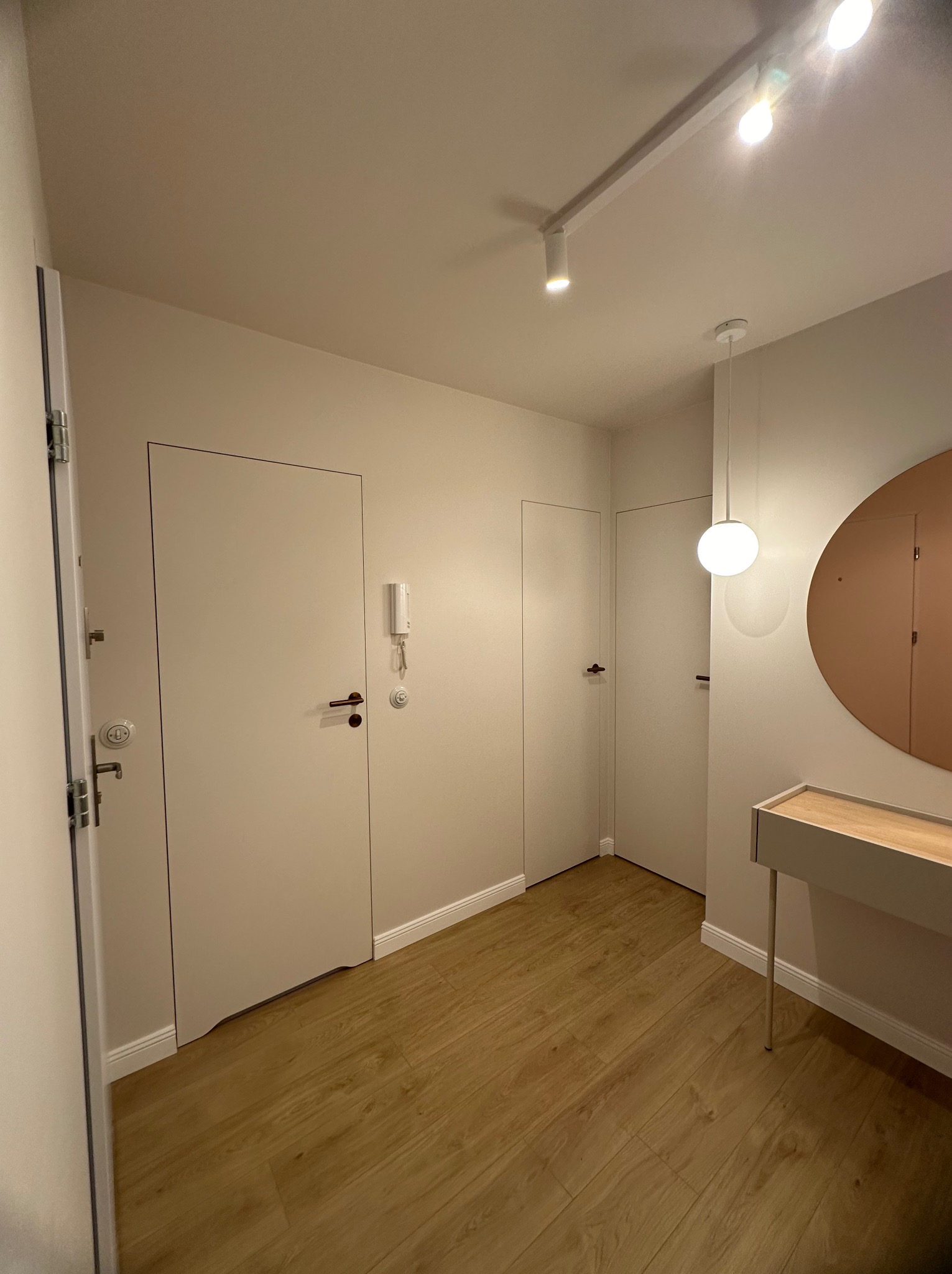 Nowoczesny przedpokój z drewnianą podłogą, białymi drzwiami ukrytymi w ścianie, okrągłym lustrem w miedzianej ramie i minimalistyczną konsolą z jasnego drewna, oświetlony reflektorami i wiszącą...