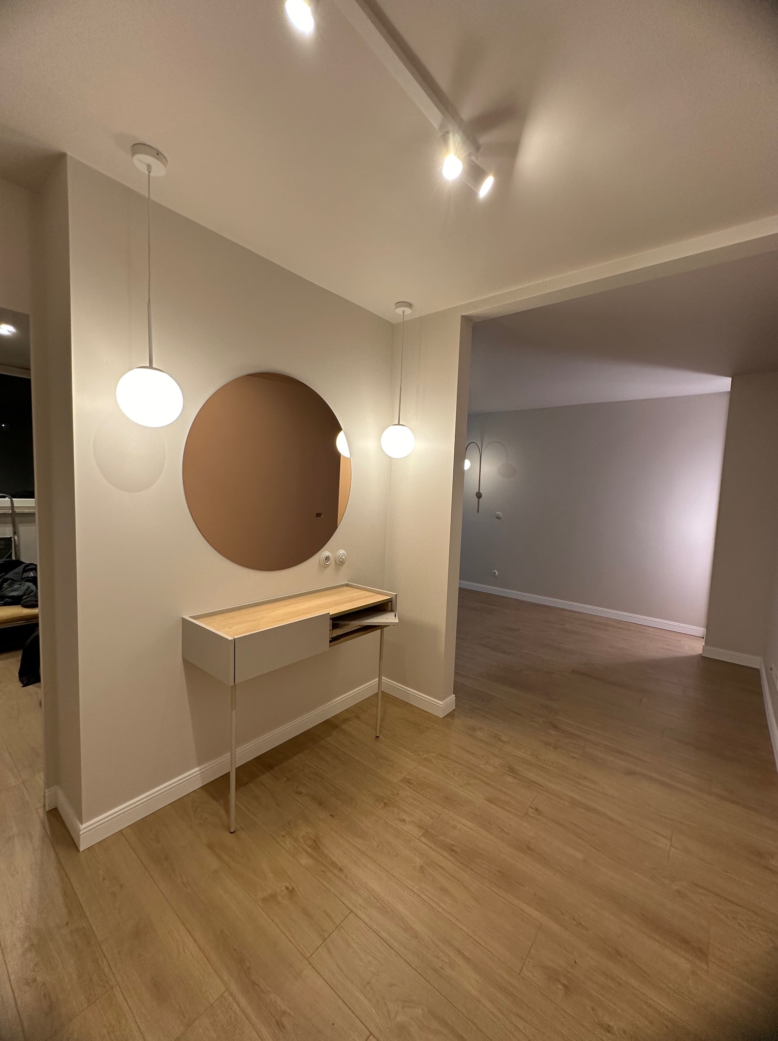 Nowoczesny przedpokój z okrągłym lustrem w miedzianej ramie, minimalistyczną konsolą z jasnego drewna i białymi, kulistymi lampami wiszącymi, oświetlony reflektorami na szynie, z podłogą wyłożoną...