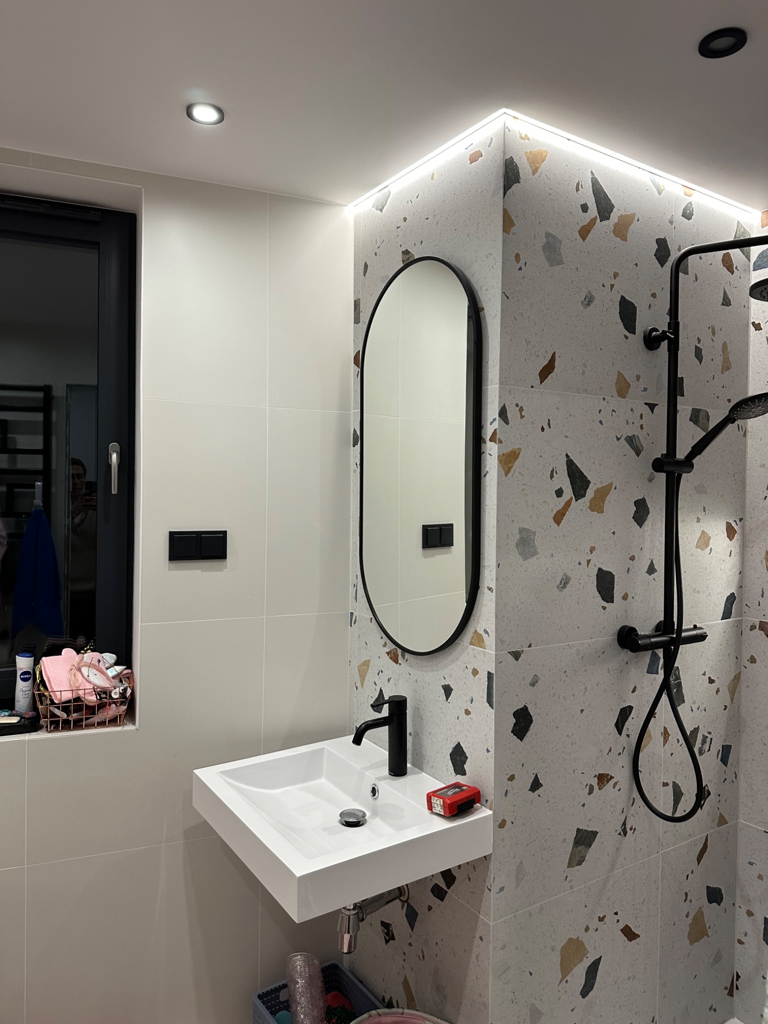 Nowoczesna łazienka po remoncie: biała, prostokątna umywalka z czarną baterią, owalne lustro w czarnej ramie, czarny zestaw prysznicowy na ścianie wyłożonej płytkami imitującymi lastryko...