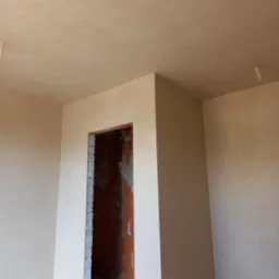 Wnętrze pokoju po nałożeniu gładzi gipsowej na ściany i sufit, widoczne niedokończone obramowanie drzwi z surowej cegły.