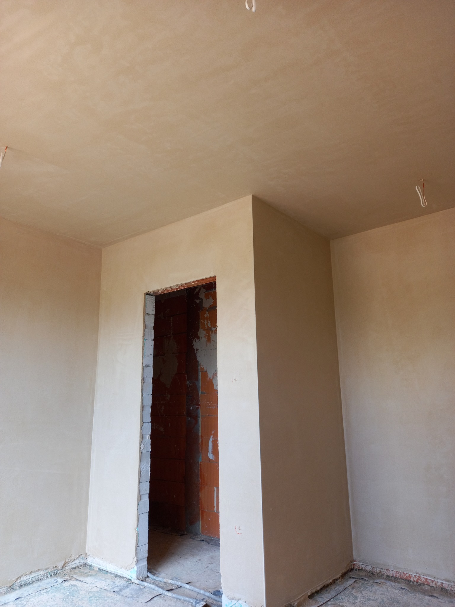 Wnętrze pokoju po nałożeniu gładzi gipsowej na ściany i sufit, widoczne niedokończone obramowanie drzwi z surowej cegły.