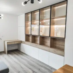 Nowoczesna zabudowa meblowa: białe szafki dolne, drewniane p&oacute;łki z czarnymi ramami i szklanymi drzwiami podświetlone LED, biurko z drewnianym blatem, szara podłoga.