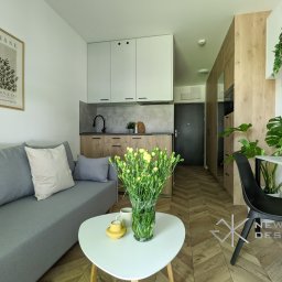 Meble New Design - Wnętrze małego mieszkania z aneksem kuchennym, szarą sofą, okrągłym białym stolikiem kawowym z bukietem żółtych kwiatów, drewnianą podłogą w jodełkę oraz biurkiem z krzesłem.