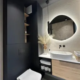 Nowoczesna łazienka z umywalką nablatową, podświetlanym lustrem o nieregularnym kształcie, czarną zabudową meblową z otwartymi półkami i wiszącą toaletą.
