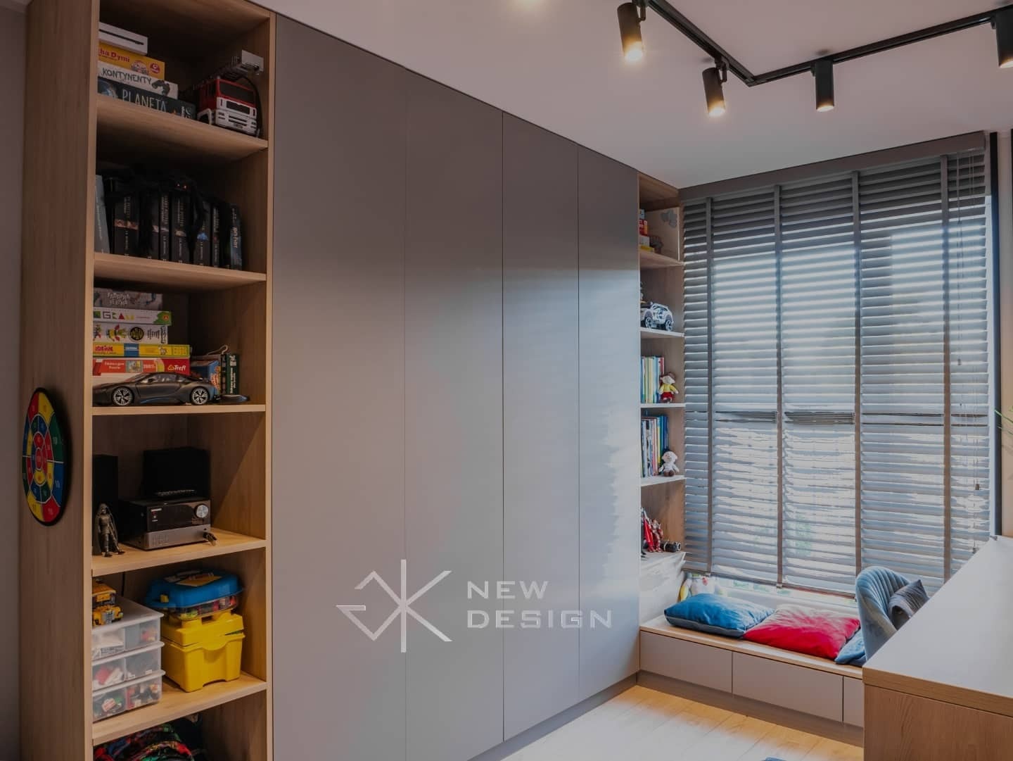 Szara zabudowa meblowa z szafą i regałami z jasnego drewna, wypełniona książkami, zabawkami i grami, oświetlona reflektorami sufitowymi i światłem z okna z żaluzjami.