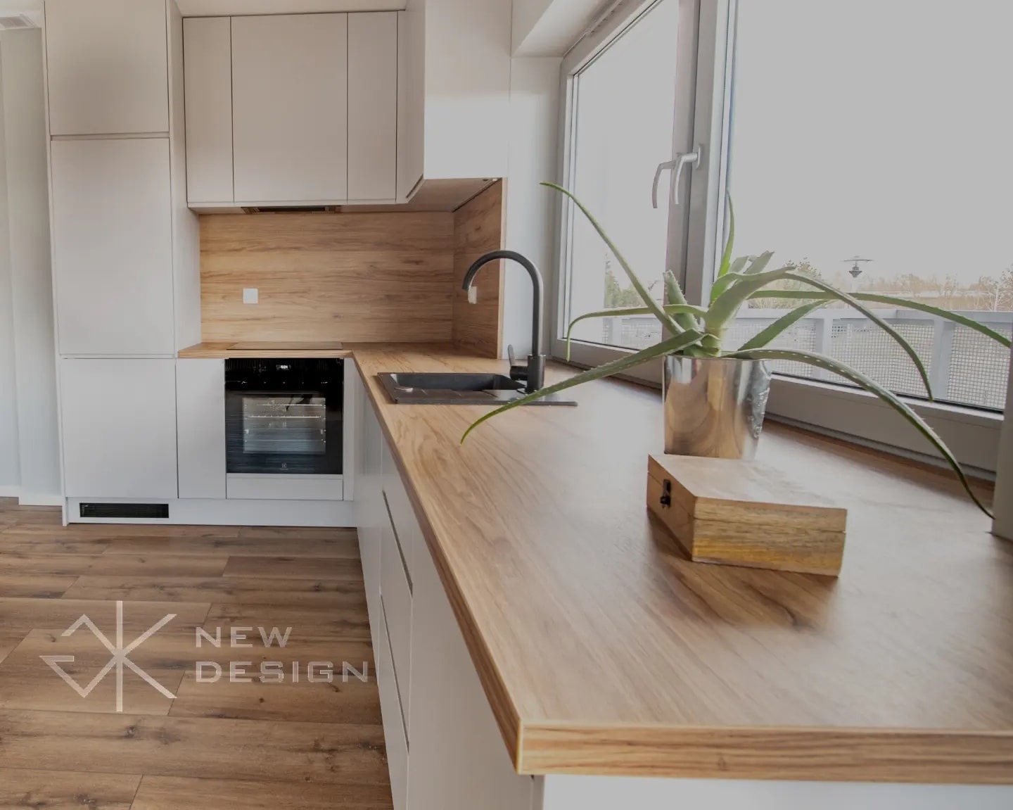 Jasna kuchnia z drewnianym blatem, czarną armaturą i rośliną w srebrnej doniczce przy oknie, widok na zewnątrz.