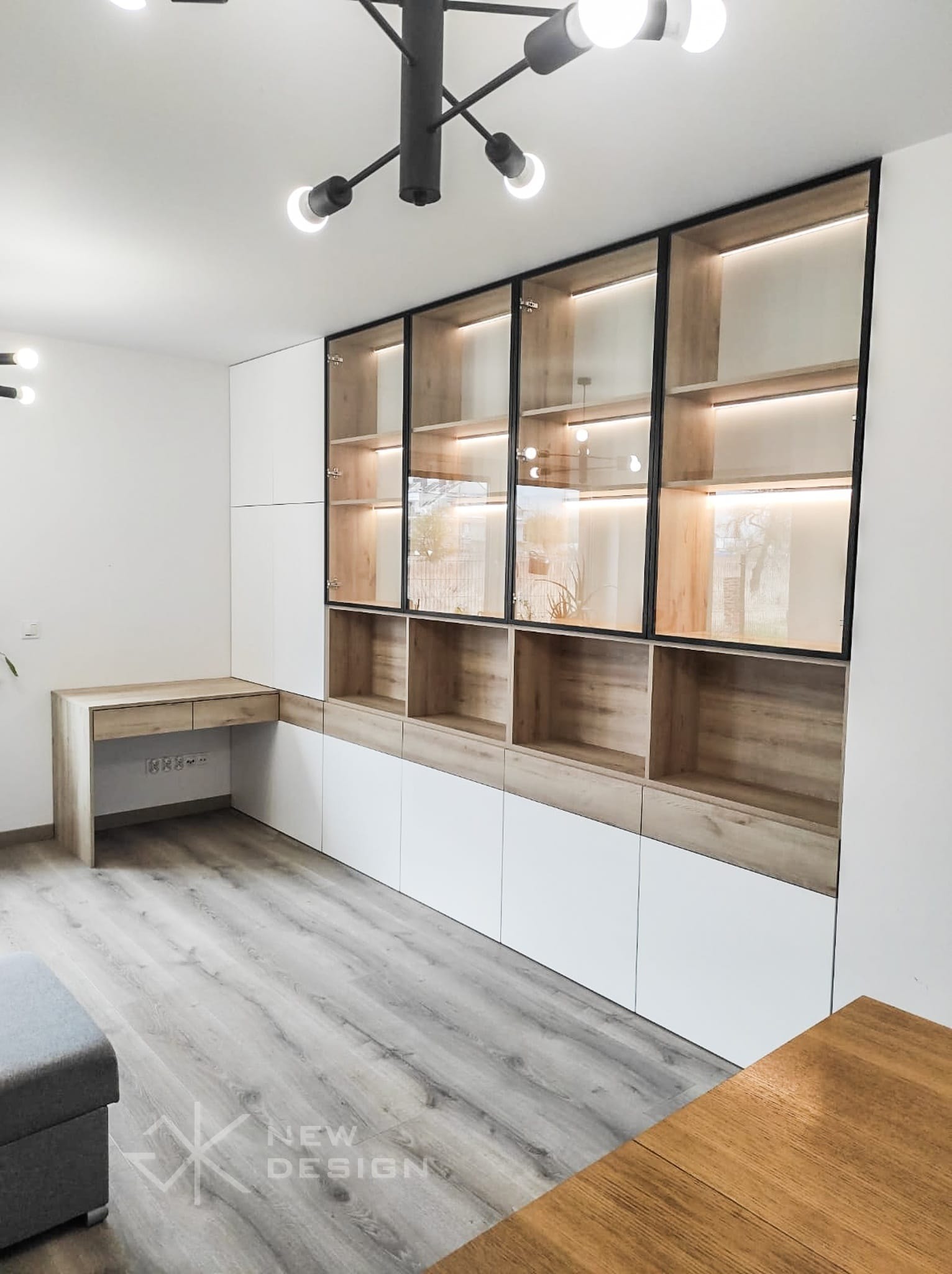Nowoczesna zabudowa meblowa: białe szafki dolne, drewniane półki z czarnymi ramami i szklanymi drzwiami podświetlone LED, biurko z drewnianym blatem, szara podłoga.