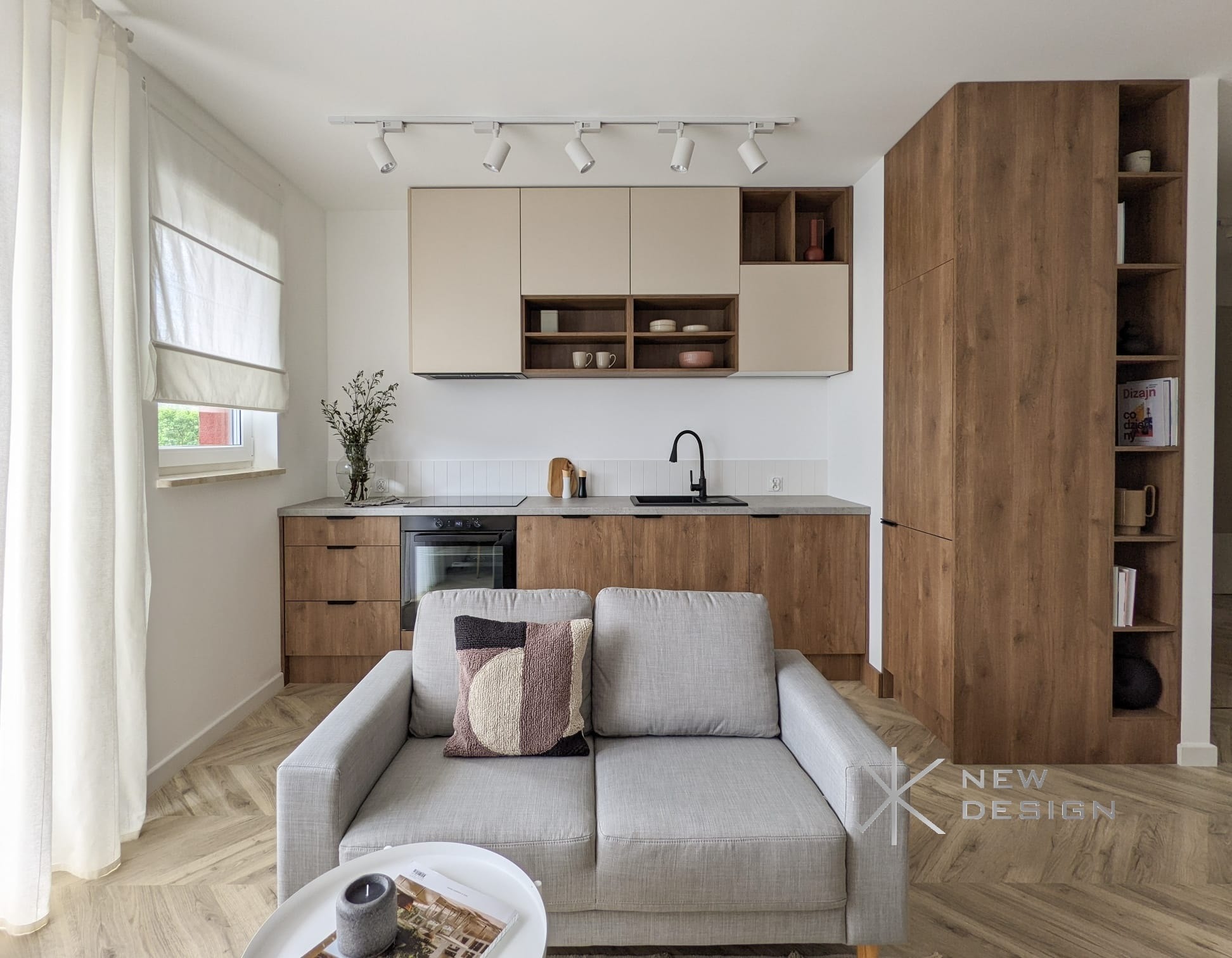 Jasna, nowoczesna kuchnia z drewnianymi i beżowymi szafkami, czarną armaturą i szarym blatem, widziana z perspektywy salonu z szarą sofą i stolikiem kawowym na parkiecie w jodełkę.