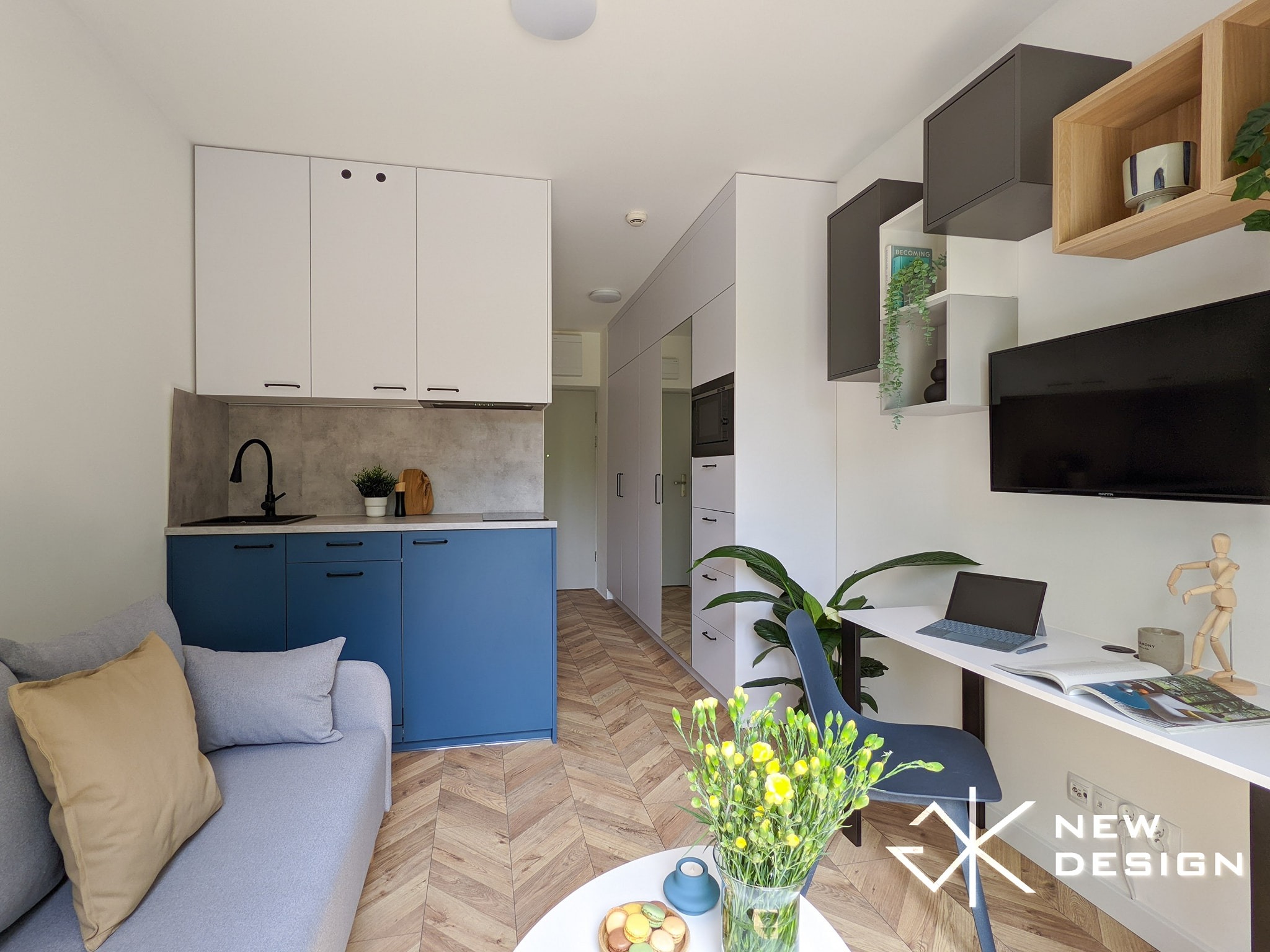 Wnętrze małego mieszkania z aneksem kuchennym: niebieskie szafki dolne, białe górne, szary blat, drewniana podłoga w jodełkę, szara sofa z poduszkami, białe biurko z laptopem i drewnianą figurką...