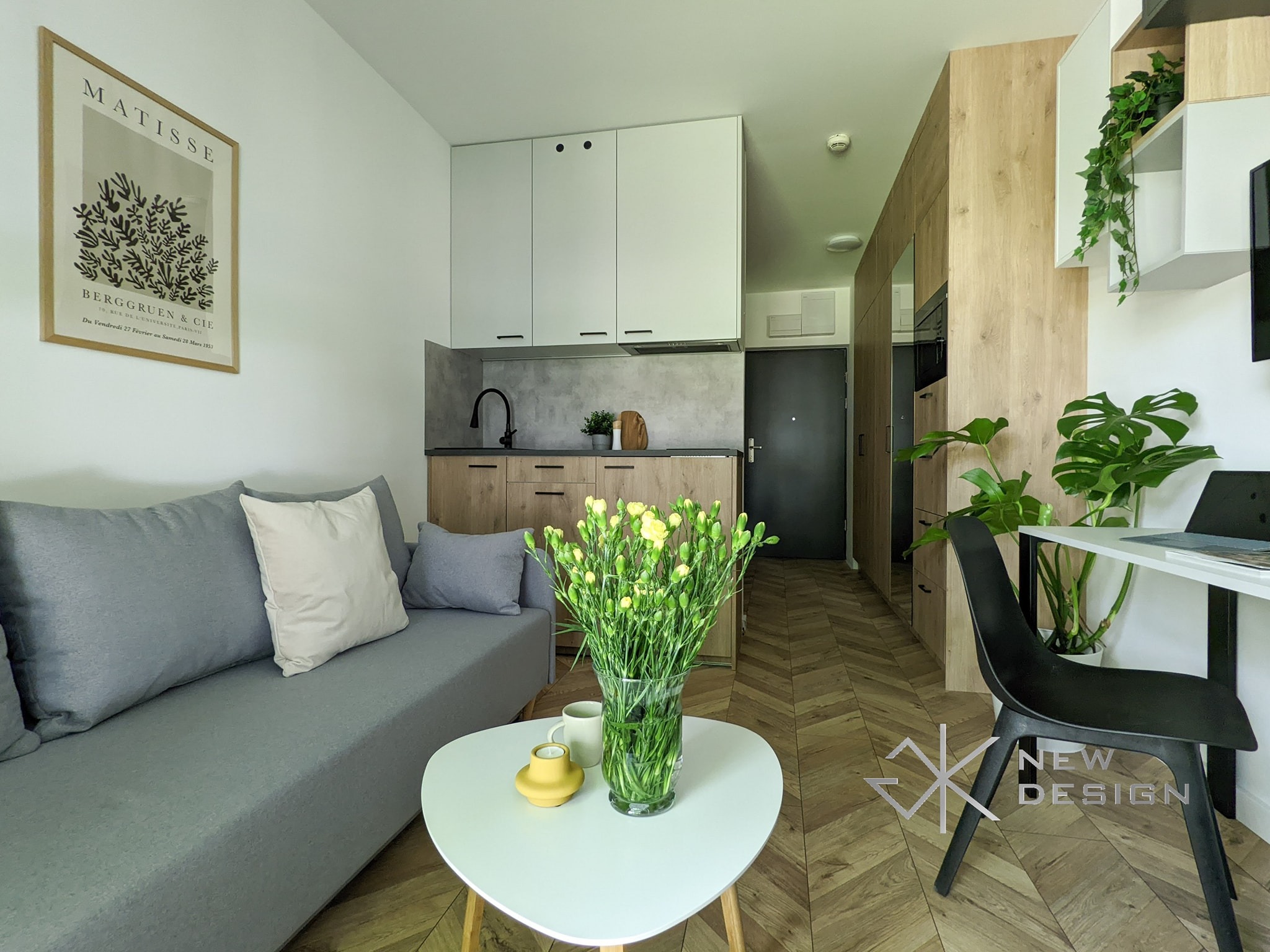 Wnętrze małego mieszkania z aneksem kuchennym, szarą sofą, okrągłym białym stolikiem kawowym z bukietem żółtych kwiatów, drewnianą podłogą w jodełkę oraz biurkiem z krzesłem.