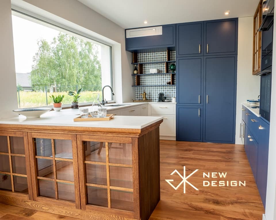 Nowoczesna kuchnia z wyspą i dużym oknem, zabudowa w odcieniach bieli i granatu, drewniana podłoga z logo na pierwszym planie.