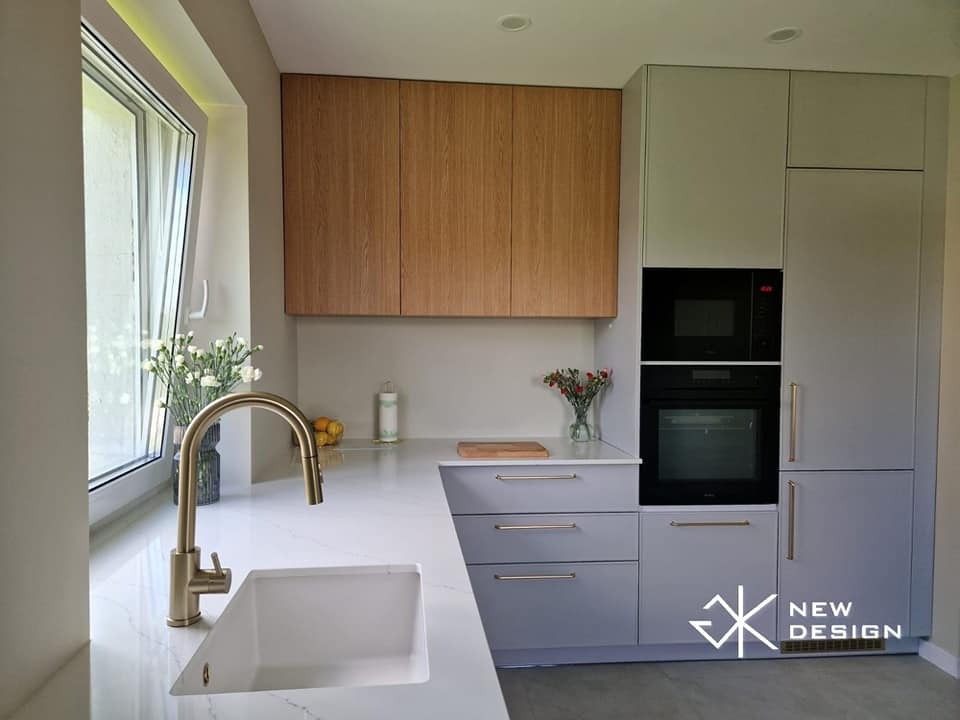 Nowoczesna kuchnia z szarymi szafkami dolnymi i drewnianymi górnymi, złota bateria przy białym zlewie, w tle okno z kwiatami w wazonie, zabudowane sprzęty AGD, logo firmy w prawym dolnym rogu.