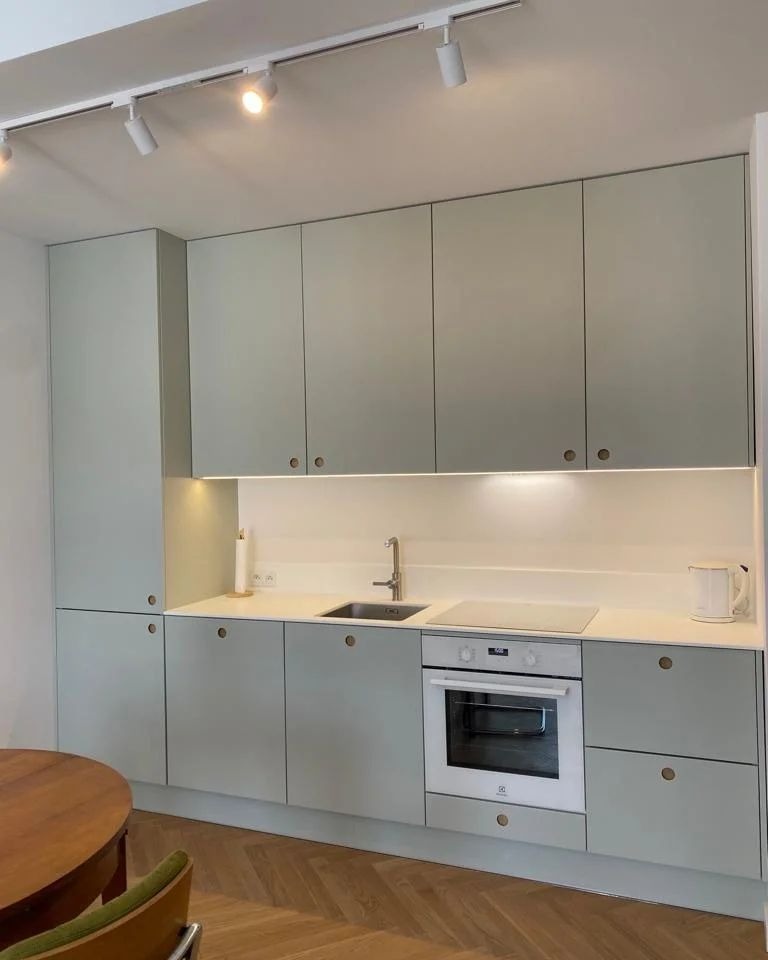 Nowoczesna kuchnia w odcieniach szarości z drewnianym blatem stołu i parkietem ułożonym w jodełkę, oświetlona listwą LED pod szafkami wiszącymi i reflektorami sufitowymi.