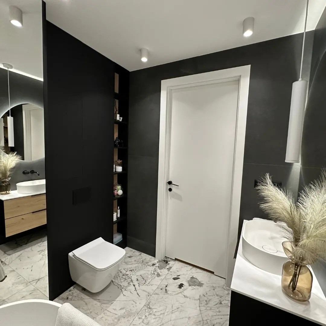 Nowoczesna łazienka w czerni i bieli z marmurową podłogą, okrągłym lustrem z podświetleniem i minimalistyczną armaturą, uzupełniona dekoracyjną trawą pampasową w szklanym wazonie.