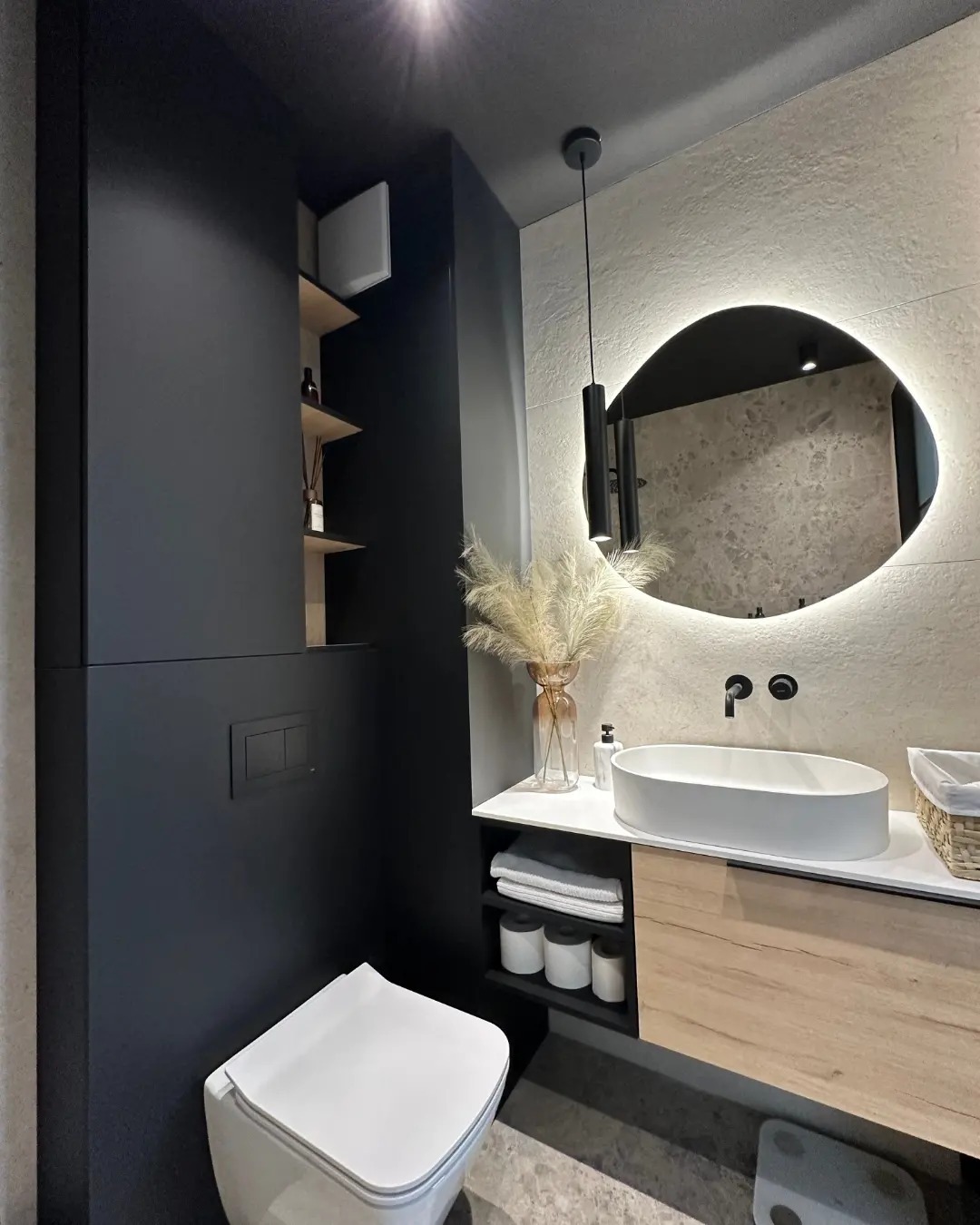 Nowoczesna łazienka z umywalką nablatową, podświetlanym lustrem o nieregularnym kształcie, czarną zabudową meblową z otwartymi półkami i wiszącą toaletą.