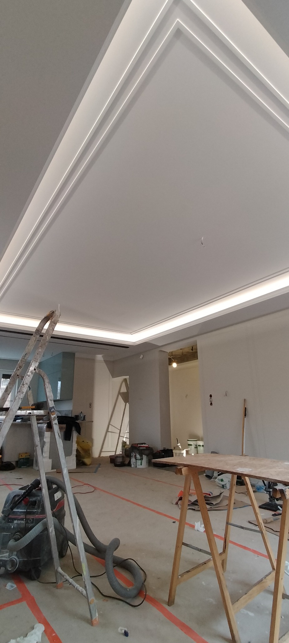 Wnętrze w trakcie remontu z widocznym sufitem z ozdobnymi listwami i oświetleniem LED, drabina i odkurzacz przemysłowy na podłodze zabezpieczonej taśmą malarską.