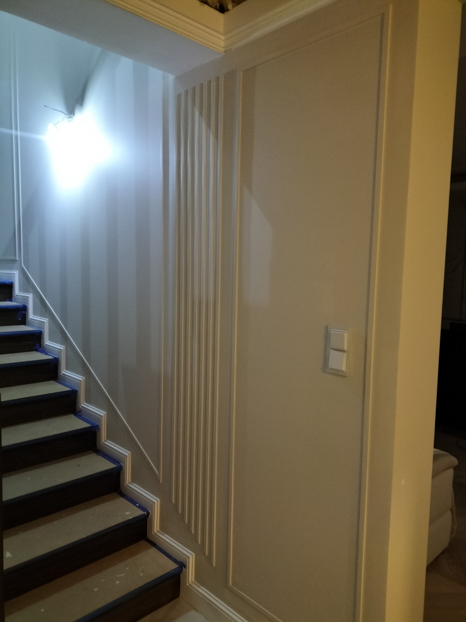 Klatka schodowa w trakcie wykończenia, zabezpieczone taśmą schody, dekoracyjne listwy pionowe na ścianie, jasne ściany i włącznik światła.