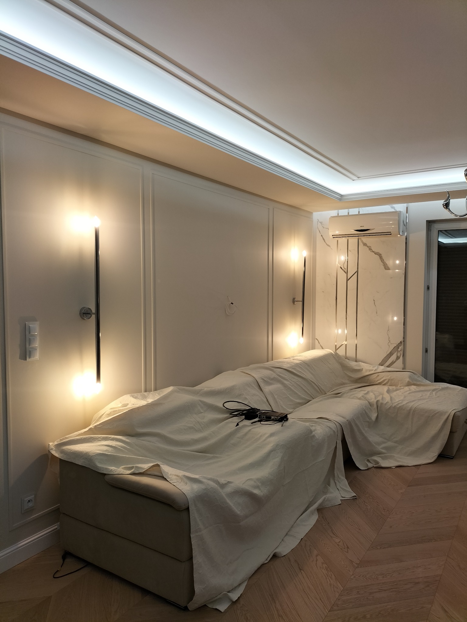 Salon w trakcie remontu: sofa przykryta płachtą ochronną, widoczne panele na ścianach, nowoczesne oświetlenie ścienne, marmurowa ściana z klimatyzatorem.