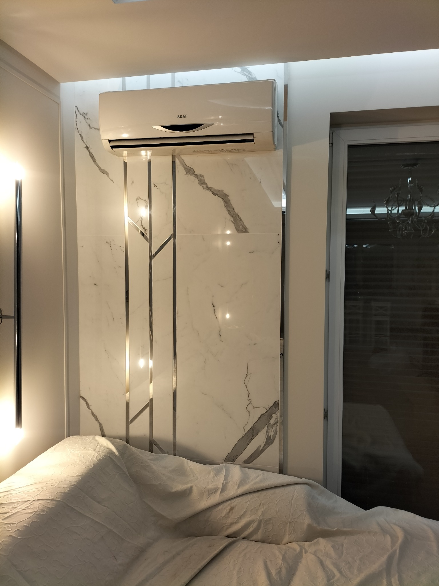 Klimatyzator marki Akai zamontowany na ścianie wyłożonej płytkami imitującymi biały marmur z metalowymi wstawkami w sypialni, widoczne okno z żyrandolem i częściowo przykryte łóżko.