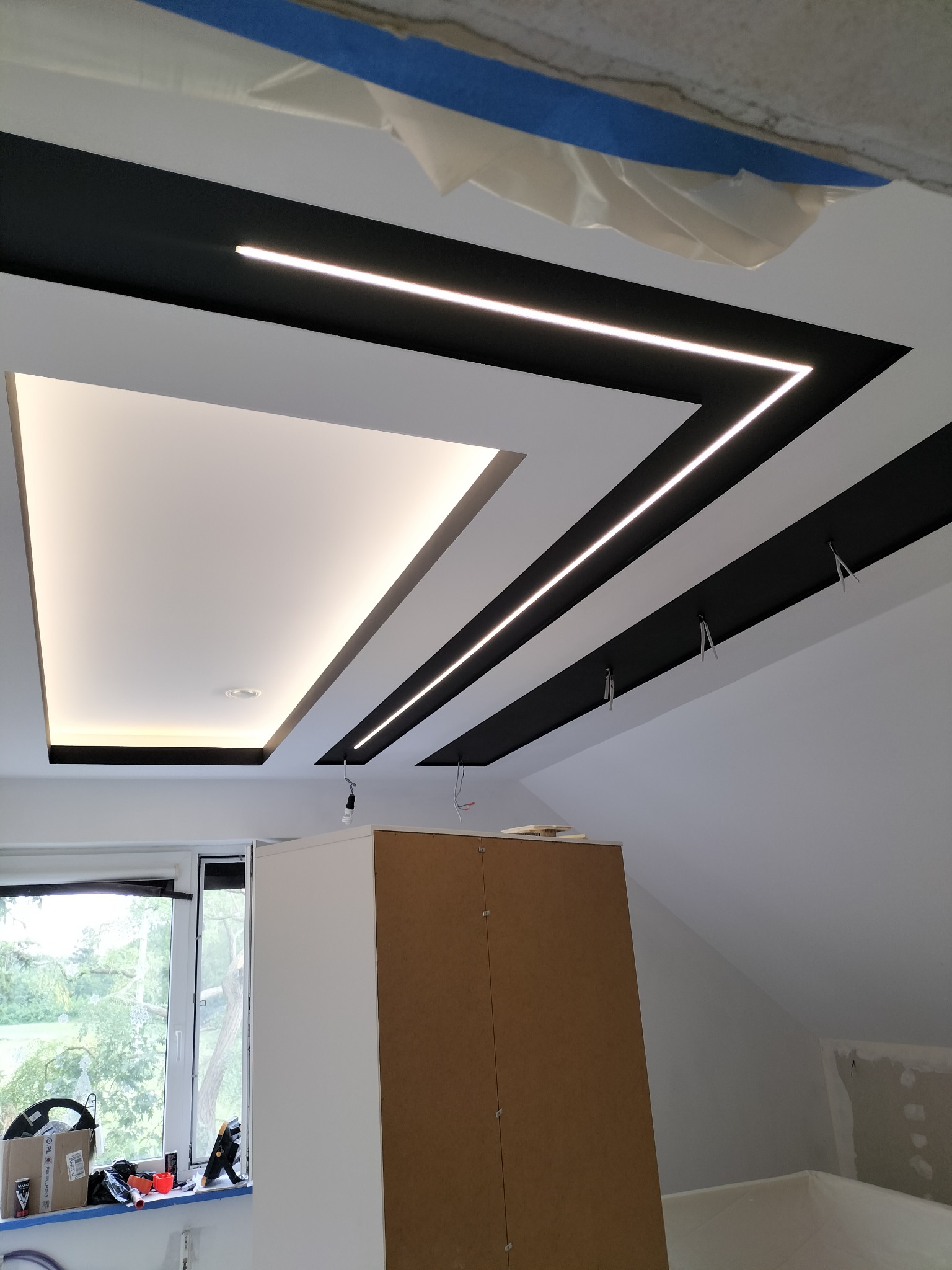 Sufit podwieszany z czarnymi elementami i oświetleniem LED w kształcie litery Z, widoczne przewody i niedokończone wykończenie ścian, częściowo zasłonięte szafką.