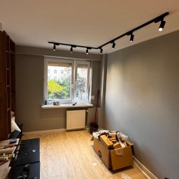 Kosztorysy i wykonawstwo Marcin Wróblewski - Pokój w trakcie remontu z zamontowanym czarnym systemem szynowym oświetlenia punktowego, kartonami na podłodze i widokiem na zieleń za oknem.