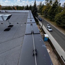 Kosztorysy i wykonawstwo Marcin Wróblewski - Instalacja odgromowa na dachu budynku z widocznymi elementami mocującymi i przewodami, tło stanowi miejski krajobraz z drzewami i ulicą.
