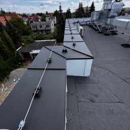 Kosztorysy i wykonawstwo Marcin Wróblewski - Widok z góry na płaski dach z zamontowaną instalacją odgromową z białym przewodem i czarnymi uchwytami, w tle zabudowa miejska i błękitne niebo z chmurami.