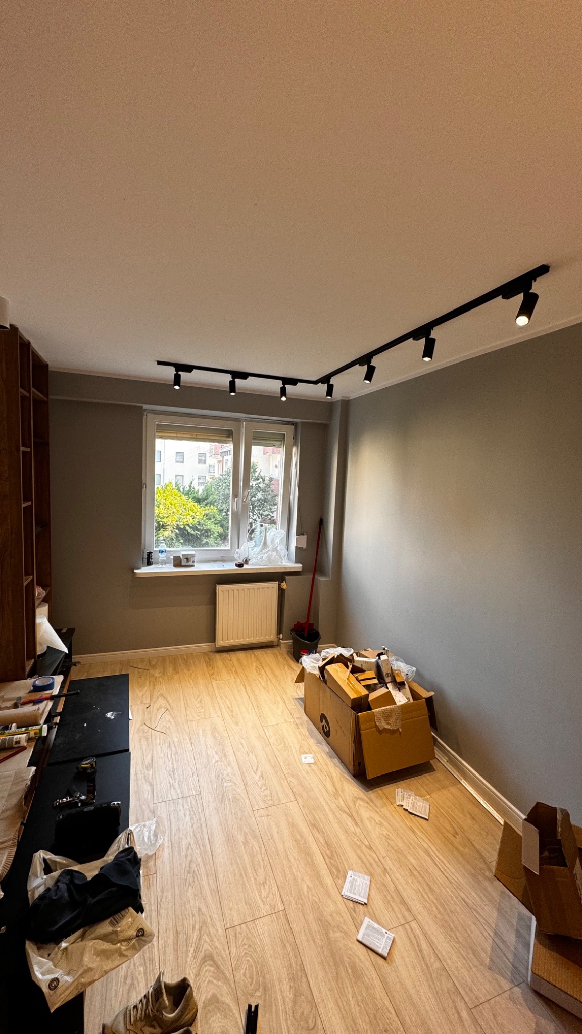 Pokój w trakcie remontu z zamontowanym czarnym systemem szynowym oświetlenia punktowego, kartonami na podłodze i widokiem na zieleń za oknem.