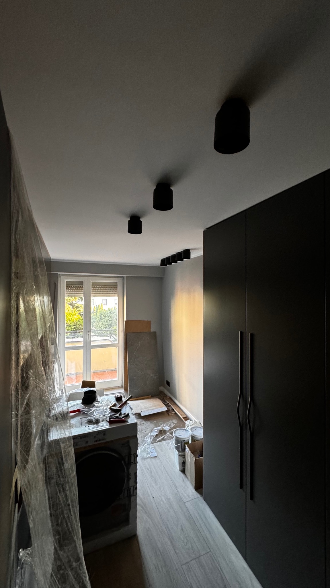 Wnętrze pokoju w trakcie remontu z widoczną pralką zabezpieczoną folią, rozłożonymi narzędziami, puszkami po farbach i zamontowanym oświetleniem sufitowym punktowym.