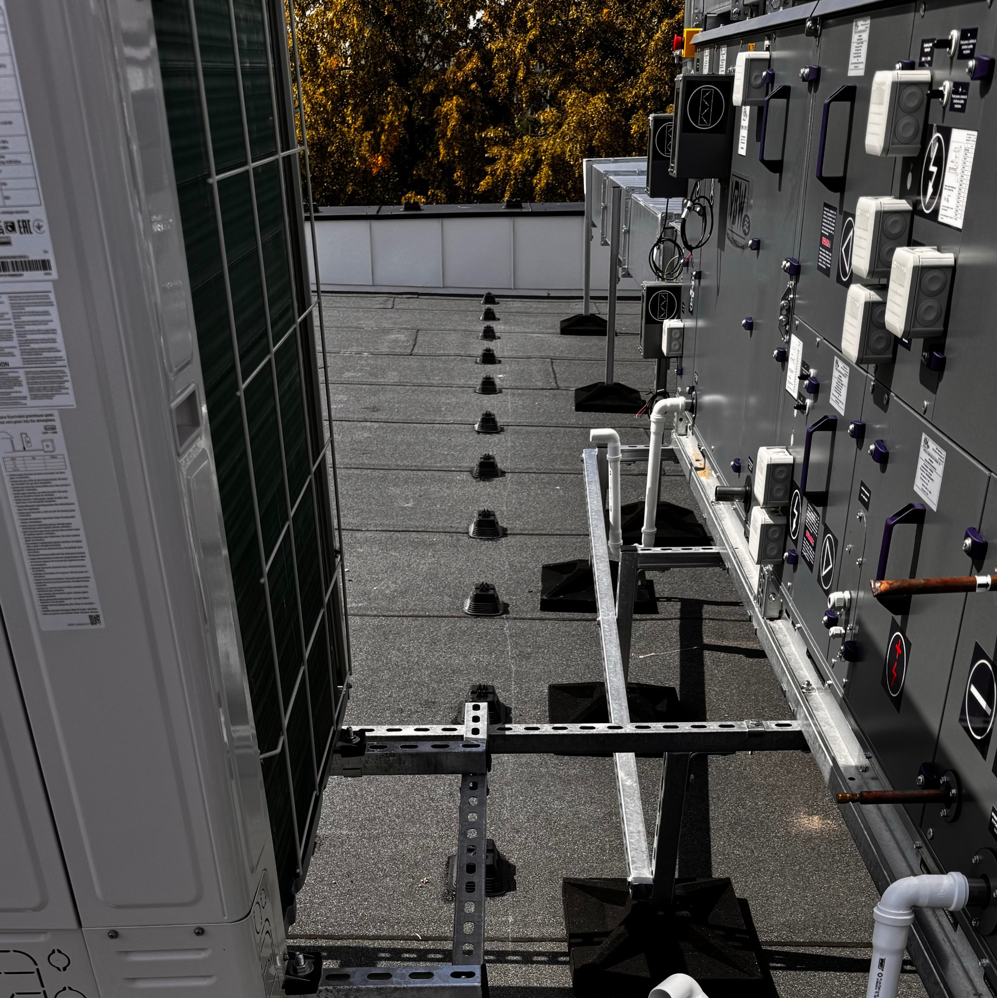 Sprzęt elektryczny na dachu budynku, w tym klimatyzator i panel sterowania z symbolami elektrycznymi i rurami, zamontowany na metalowej konstrukcji wsporczej.