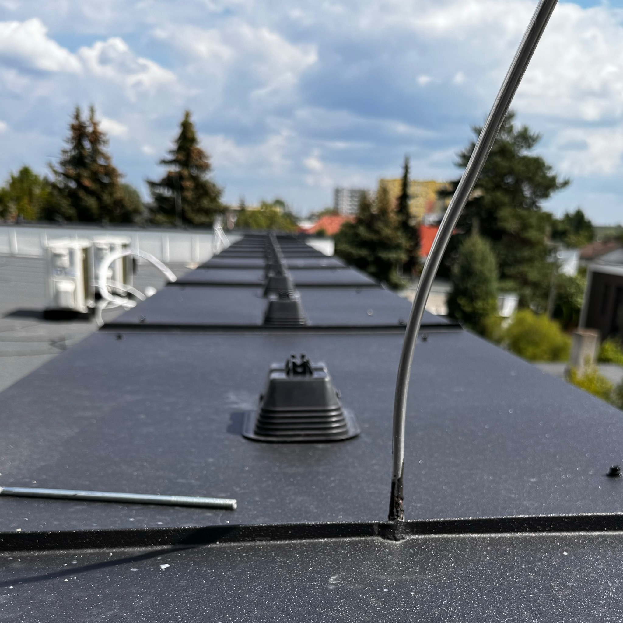 Szereg czarnych podstaw instalacji odgromowej na płaskim dachu, z widocznym przewodem odprowadzającym i elementami montażowymi, na tle błękitnego nieba z chmurami i zieleni drzew.