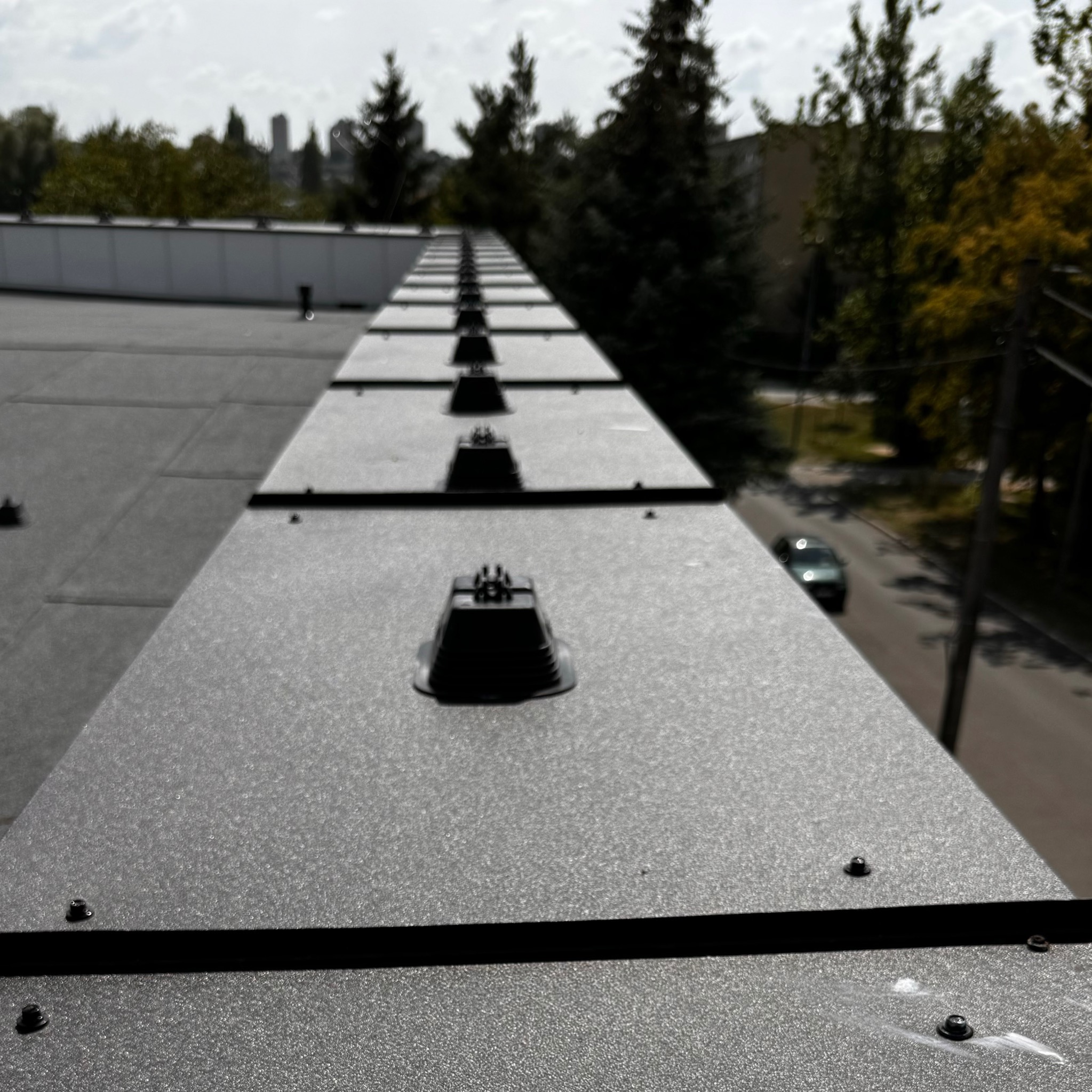 Szary dach z zamontowanymi, czarnymi uchwytami odgromowymi w rzędzie, widok z góry na ulicę i drzewa w tle.