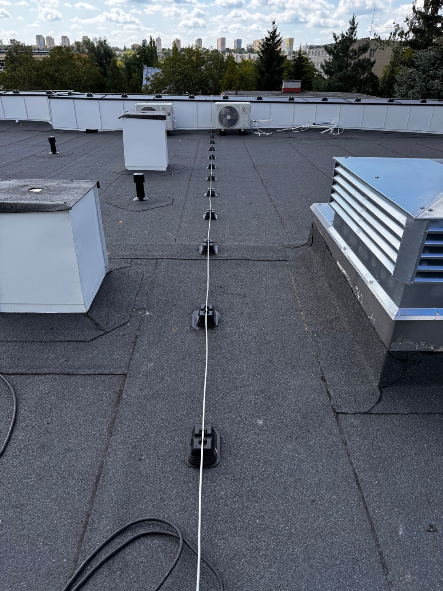 Przewód odgromowy poprowadzony po płaskim dachu pokrytym papą, z widocznymi uchwytami mocującymi, klimatyzatorami i wentylacją w tle zabudowa miejska.