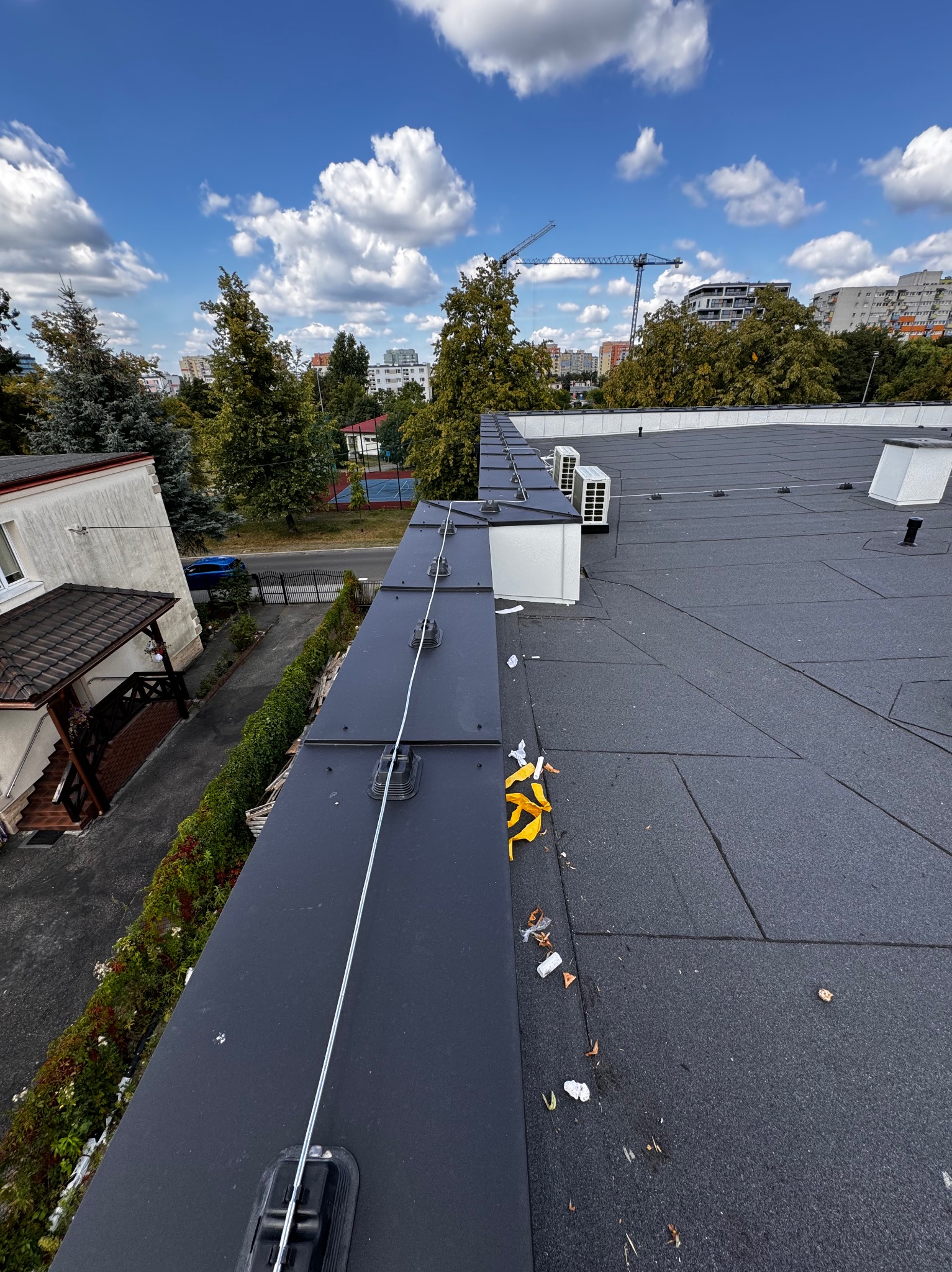 Widok z dachu budynku w Inowrocławiu na zainstalowaną instalację odgromową z metalowymi uchwytami i przewodami, w tle budynki mieszkalne i żurawie budowlane.