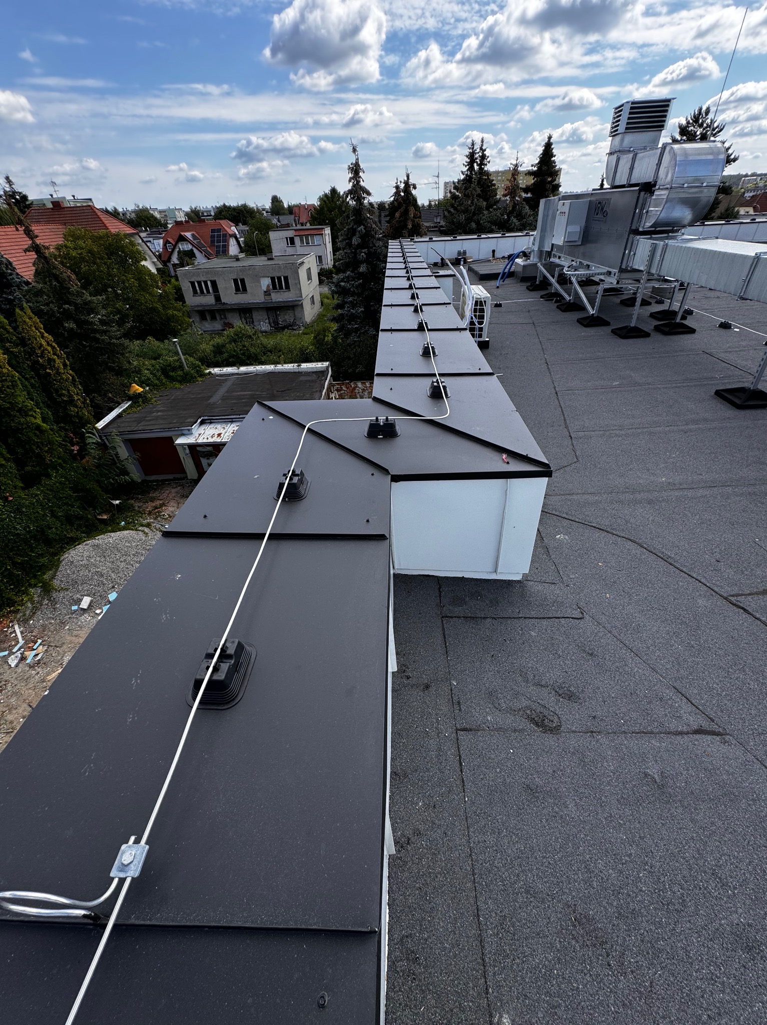 Widok z góry na płaski dach z zamontowaną instalacją odgromową z białym przewodem i czarnymi uchwytami, w tle zabudowa miejska i błękitne niebo z chmurami.