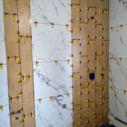 PCmedia24 Szczepan Jasiński - Układanie płytek imitujących drewno i marmur na ścianie z użyciem klipsów poziomujących. Widoczne otwory na instalacje i fragment podłogi z ogrzewaniem.