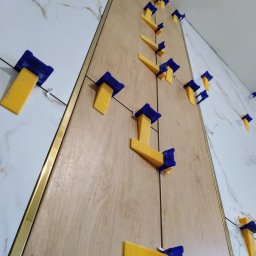 PCmedia24 Szczepan Jasiński - Układanie płytek imitujących drewno i marmur z użyciem systemu poziomowania. Widoczne żółto-niebieskie klipsy i złota listwa wykończeniowa.