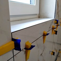 PCmedia24 Szczepan Jasiński - Układanie płytek marmuropodobnych przy oknie, z użyciem żółto-niebieskich klinów poziomujących. Widoczne krzyżyki dystansowe i fragment okna.