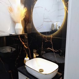 PCmedia24 Szczepan Jasiński - Elegancka łazienka z czarnymi płytkami imitującymi marmur, owalną umywalką nablatową z złotymi akcentami i okrągłym lustrem z podświetleniem LED.