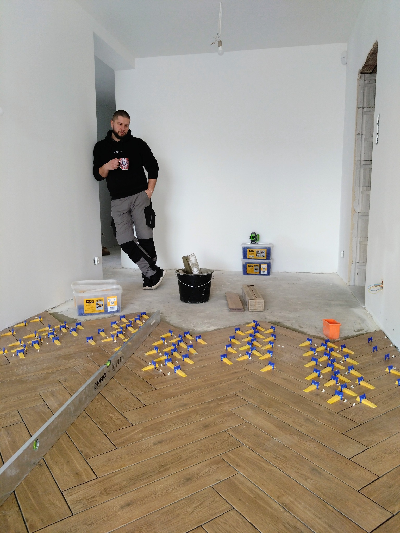 Układanie parkietu w jodełkę z użyciem klipsów poziomujących. Mężczyzna z kubkiem obserwuje postęp prac. Widoczne narzędzia i materiały budowlane.