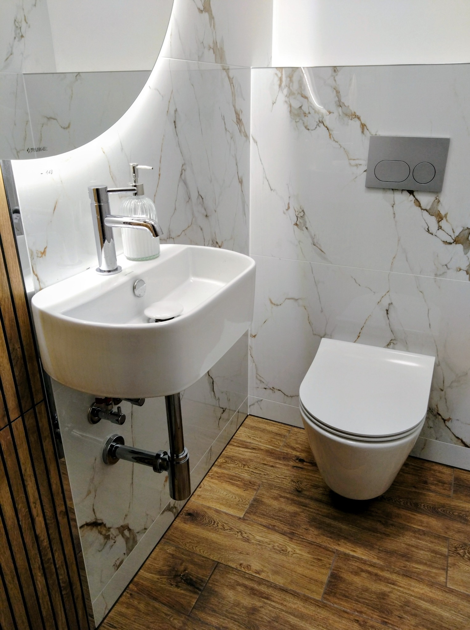 Nowoczesna łazienka z marmurowymi płytkami, drewnopodobną podłogą i podświetlanym lustrem. Umywalka nablatowa i podwieszana toaleta.
